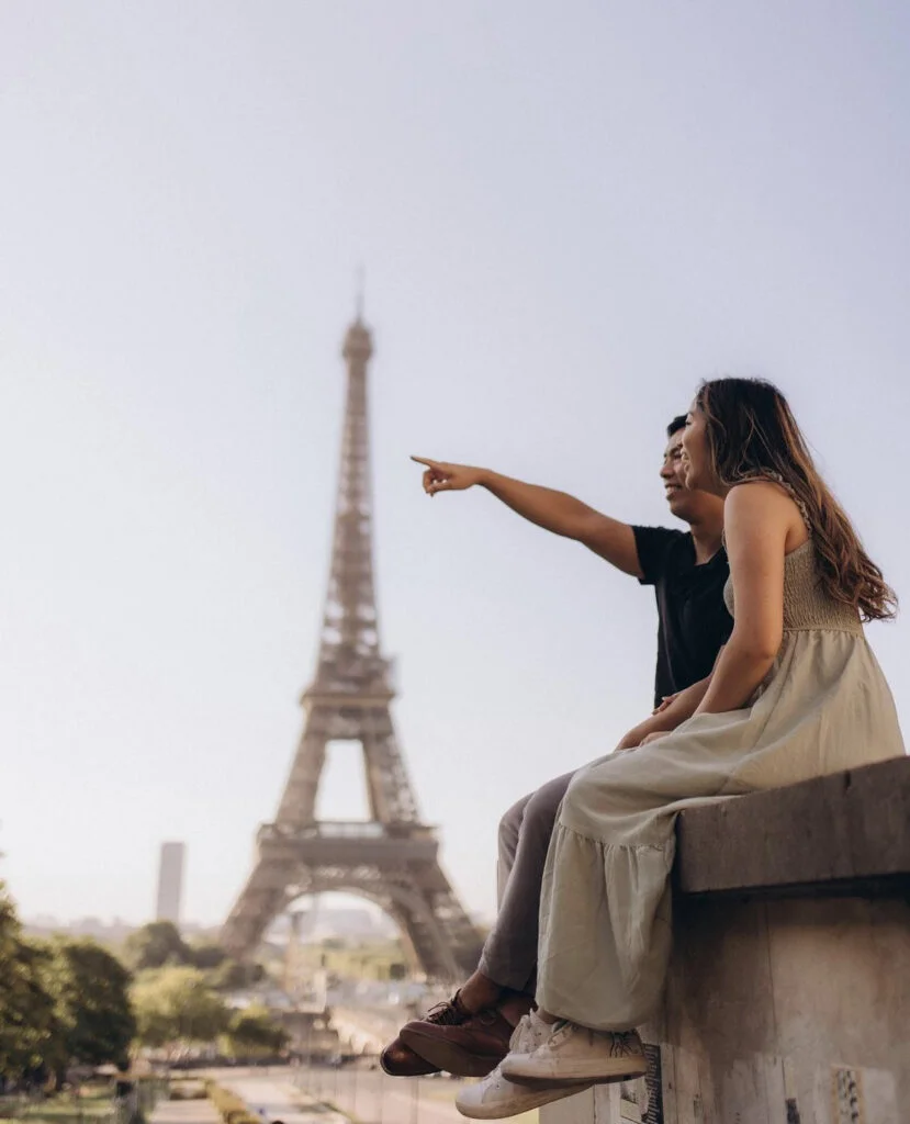 A couple sitting on a ledge at Trocadero while one points toward the tower during a Paris Eiffel Tower photoshoot