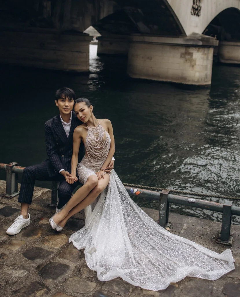 Elegant newly engaged couple posing by the Seine beneath a stone bridge during a Paris engagement photoshoot with Julia Litvin, the bride-to-be in a shimmering beaded gown with a long train