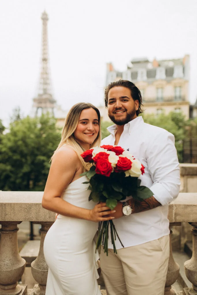A couple holding a bouquet of red and white roses on a balcony with the tower behind them during an Eiffel Tower photoshoot in Paris