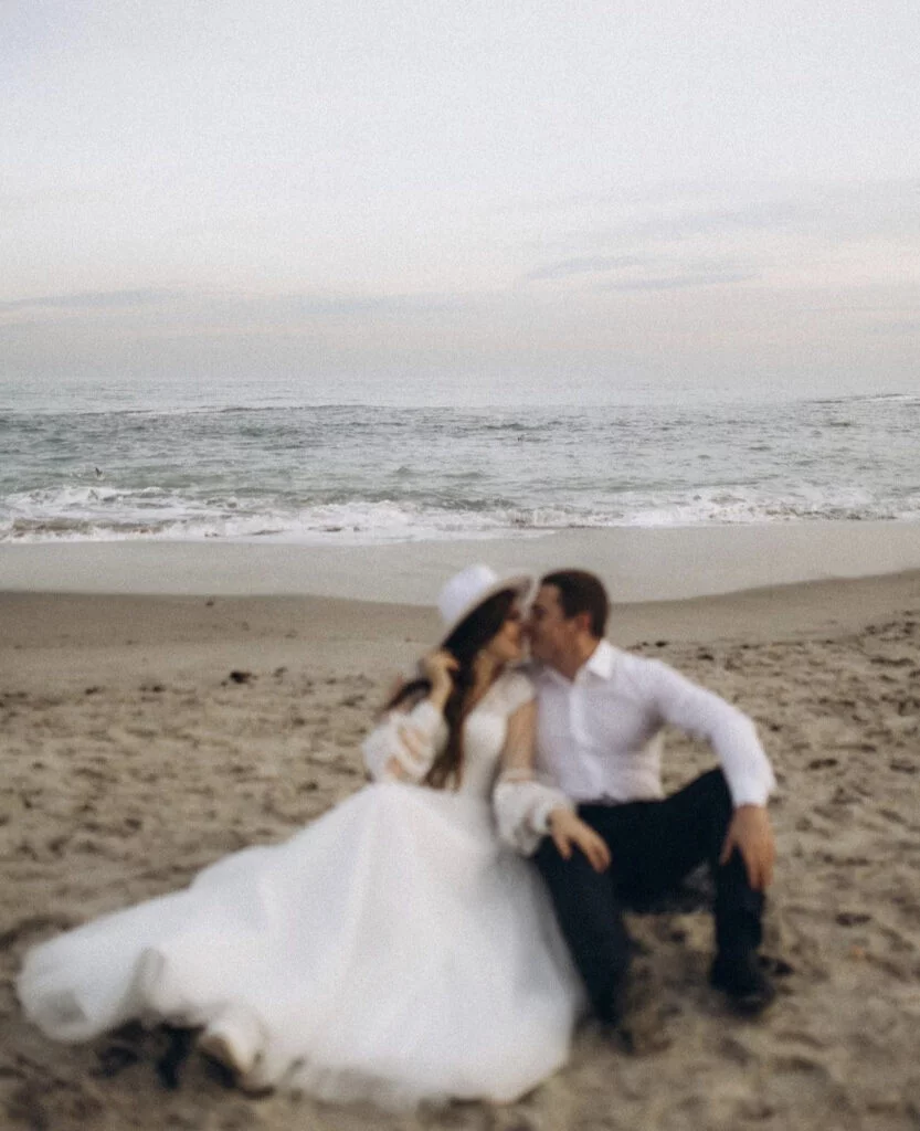 Newly engaged couple sharing a kiss on a sandy beach at dusk during a seaside engagement photoshoot with Julia Litvin, the bride-to-be in a flowing white gown and hat
