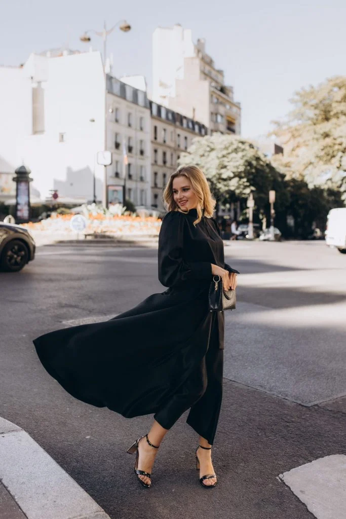 A long black dress swirling mid-turn on a sunny Parisian square, framed by pale facades and blossoming trees.