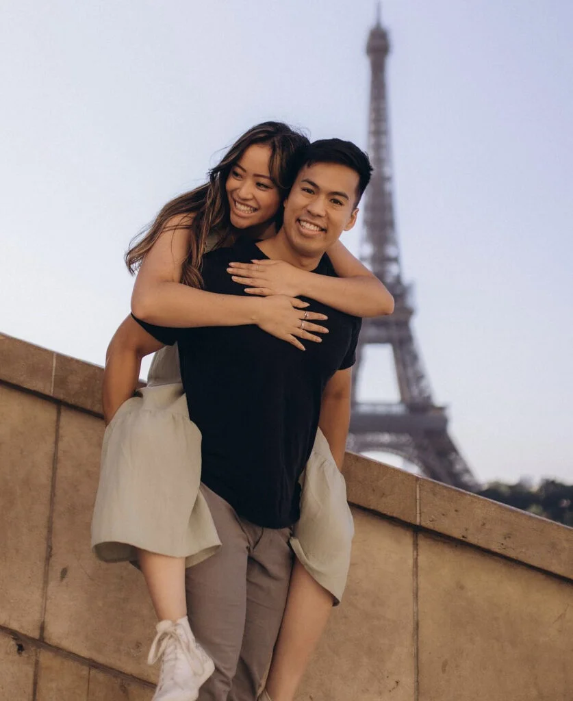 A smiling couple sharing a piggyback ride during an Eiffel Tower photoshoot in Paris on a sunny day