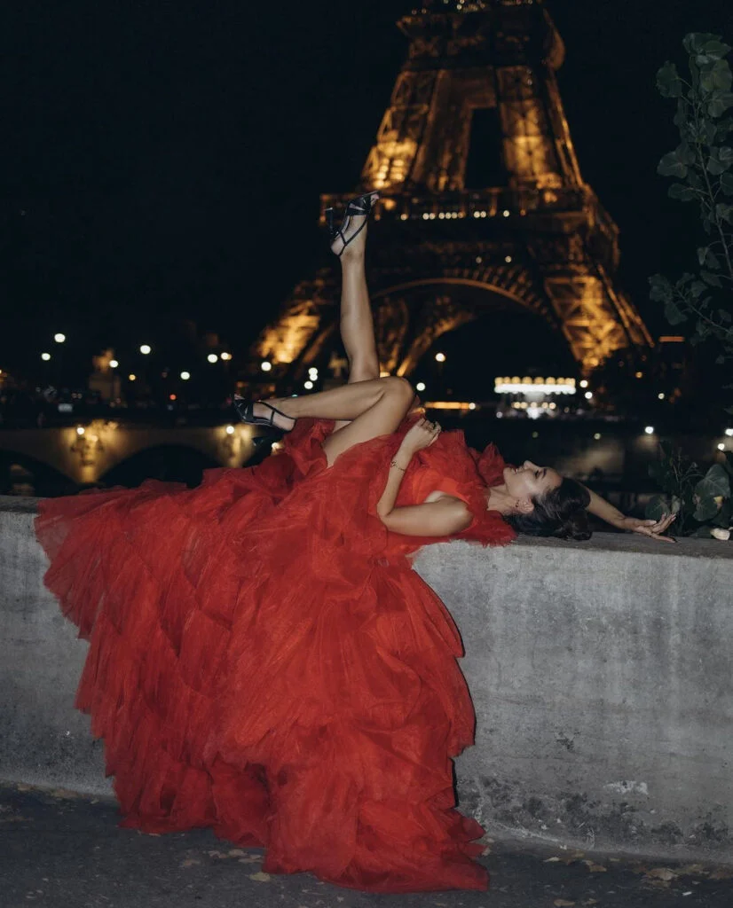 Bold night portrait photography in Paris, a woman reclining in a red gown with her legs lifted, the Eiffel Tower glowing behind