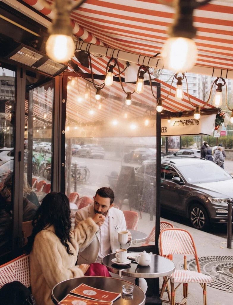 Man tenderly kisses his partner's hand across a small table at a warm Parisian sidewalk cafe