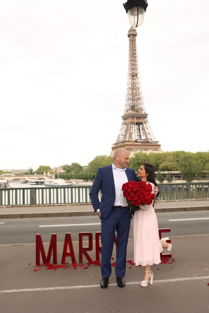 Newly engaged couple embrace with red roses beside Marry Me letters and the Eiffel Tower