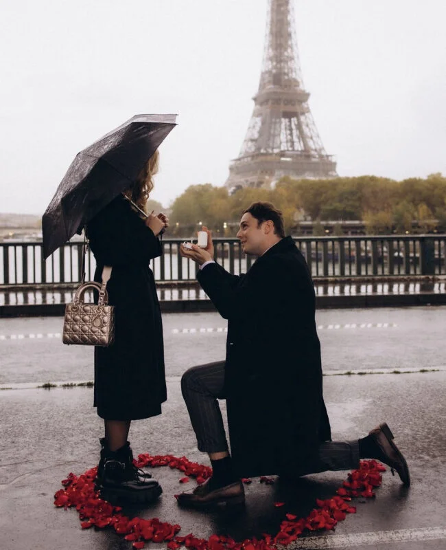 Man kneels to propose in the rain inside a heart of rose petals as his partner holds an umbrella