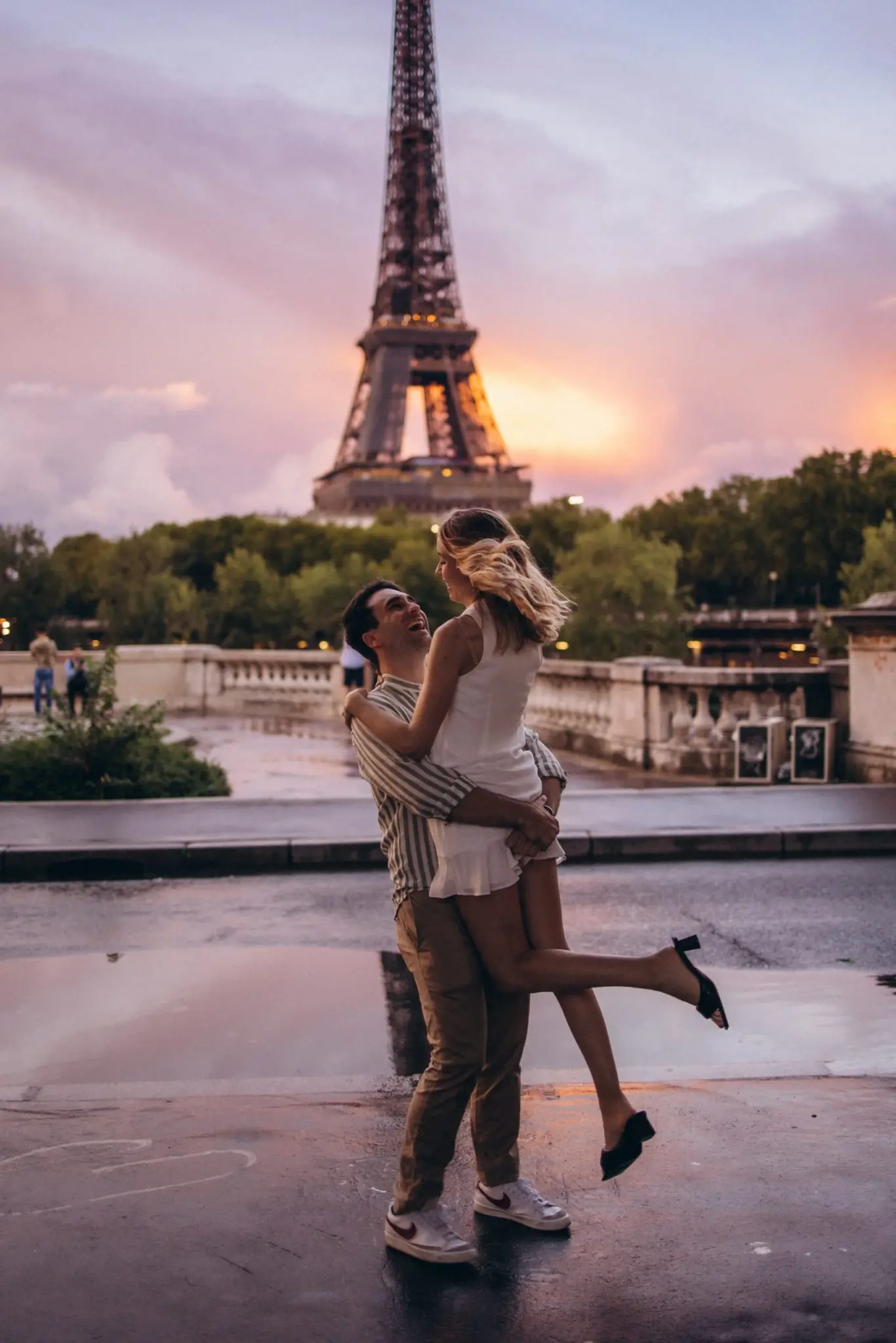 Sunset Eiffel Tower photoshoot of a couple, the man lifting his laughing partner on the wet stone of Trocadéro as the sky glows pink behind the tower