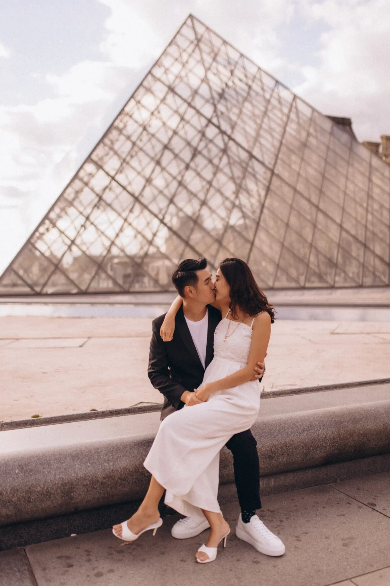 Couple photoshoot at the Louvre in Paris, a couple kissing while seated on the stone rim of the fountain in front of the glass pyramid