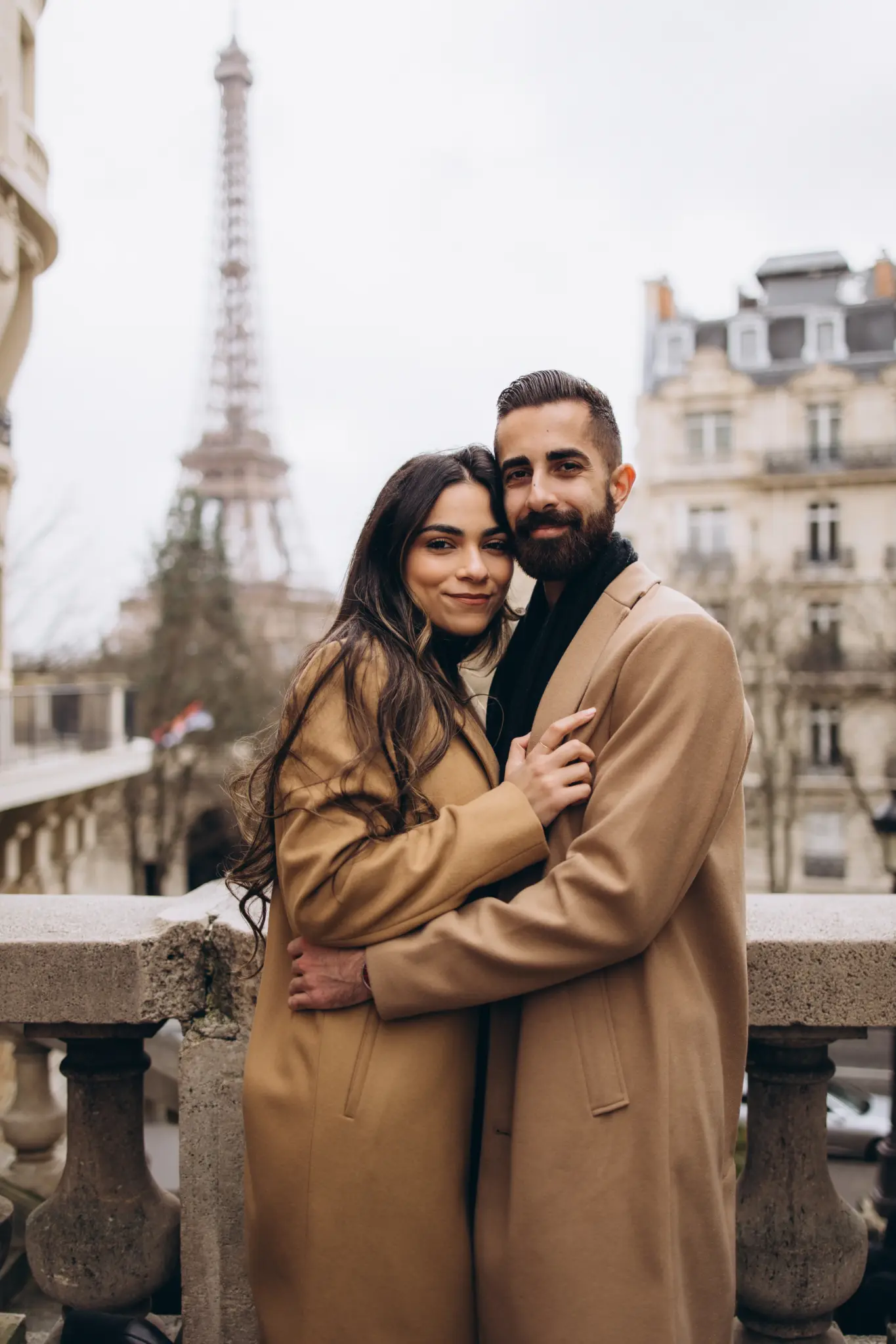 Couple in camel coats at the stone balustrade of Avenue de Camoëns with the Eiffel Tower behind them