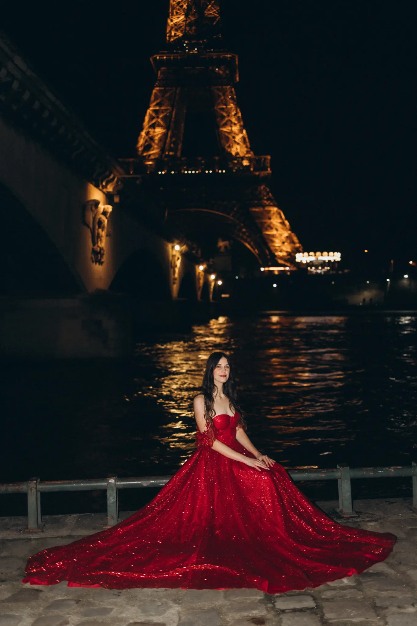 Woman in a red gown sitting on the Seine quay at night with the Eiffel Tower lit up