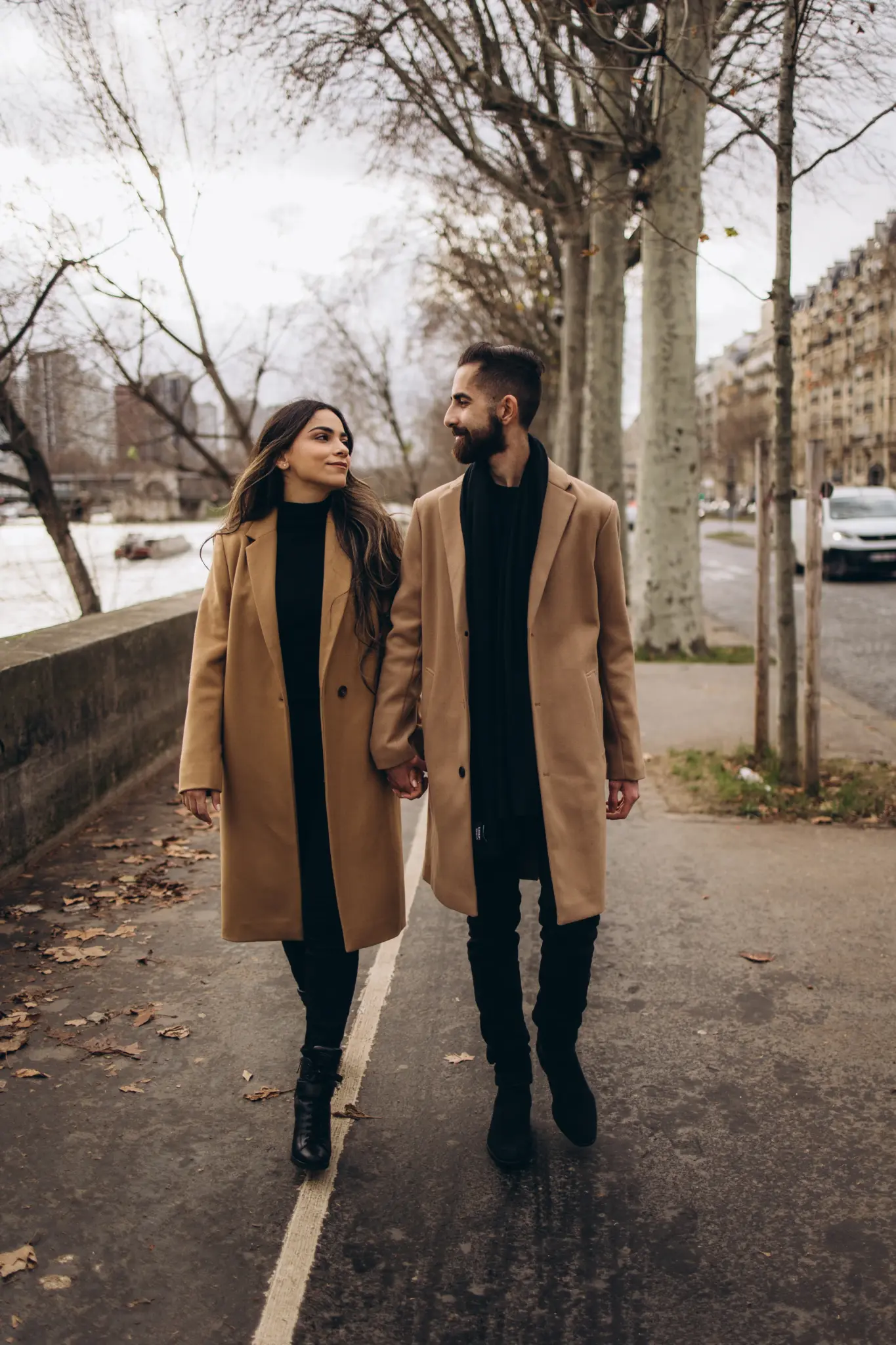 Couple walking hand in hand along a tree-lined quay near the Eiffel Tower