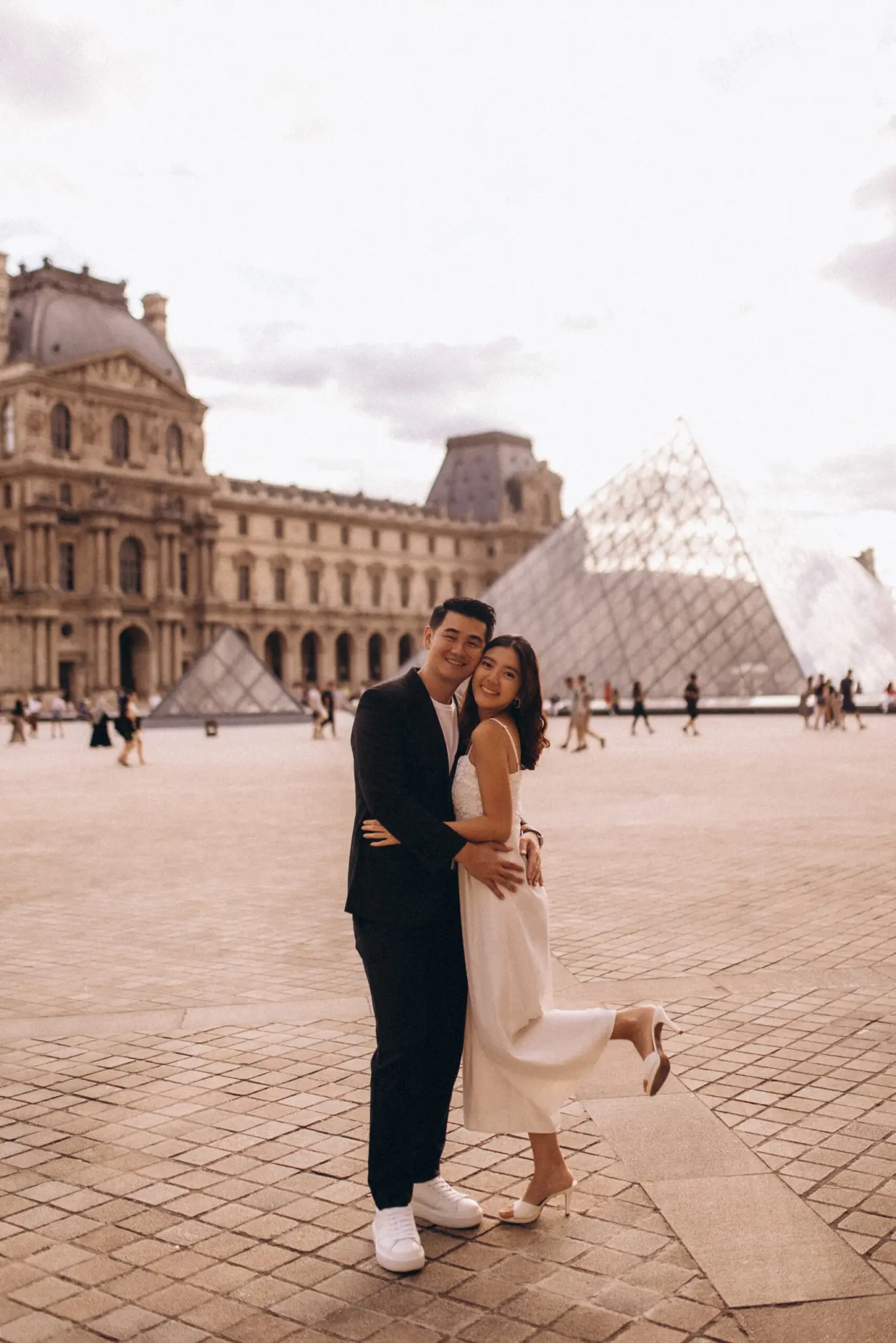 Louvre couple photoshoot in Paris, a couple embracing and laughing in front of the Louvre palace and its glass pyramid, the woman lifting one heel