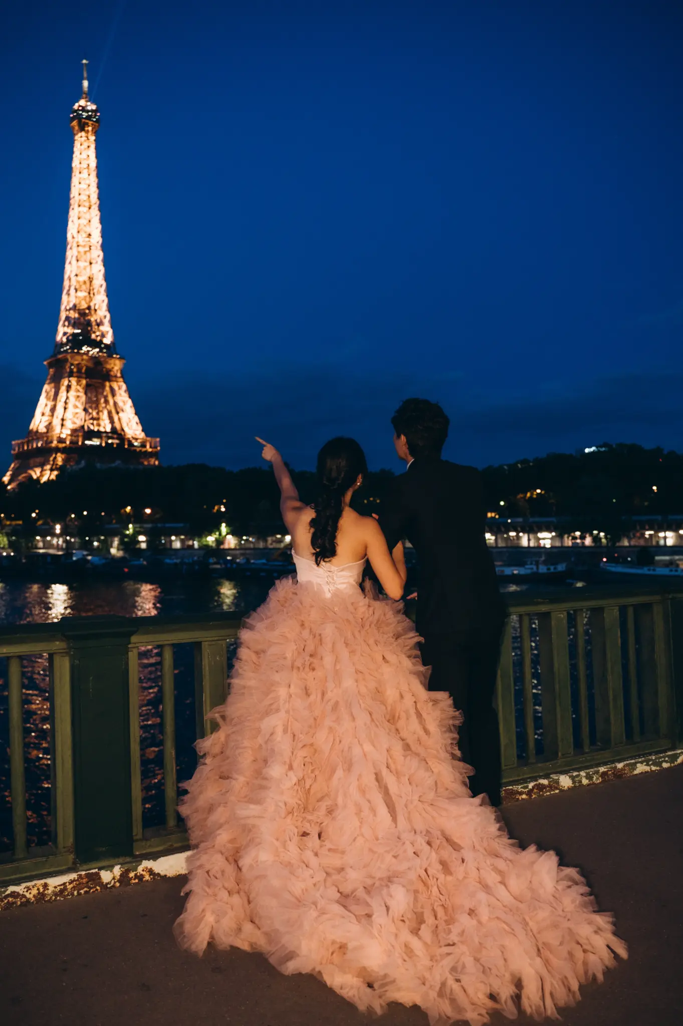 Woman in a pink tulle gown with her partner at night facing the lit Eiffel Tower