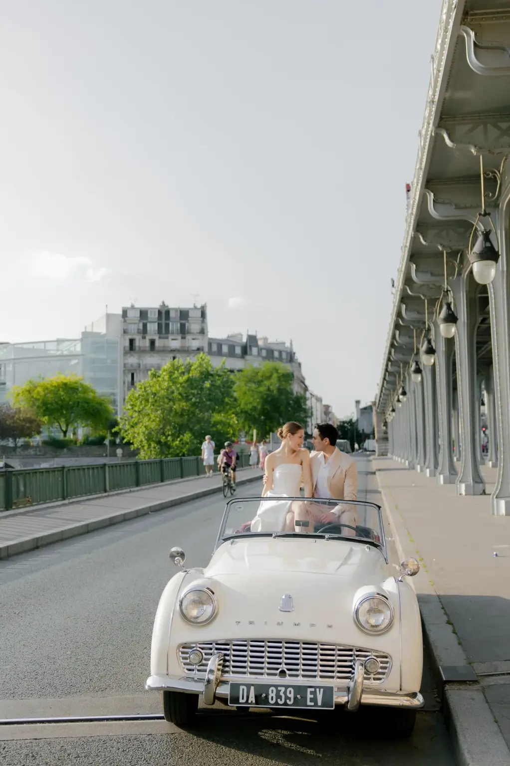 Paris couple photoshoot in a vintage white Triumph convertible on the Bir-Hakeim bridge, a couple in a pale gown and beige suit, by Julia Litvin