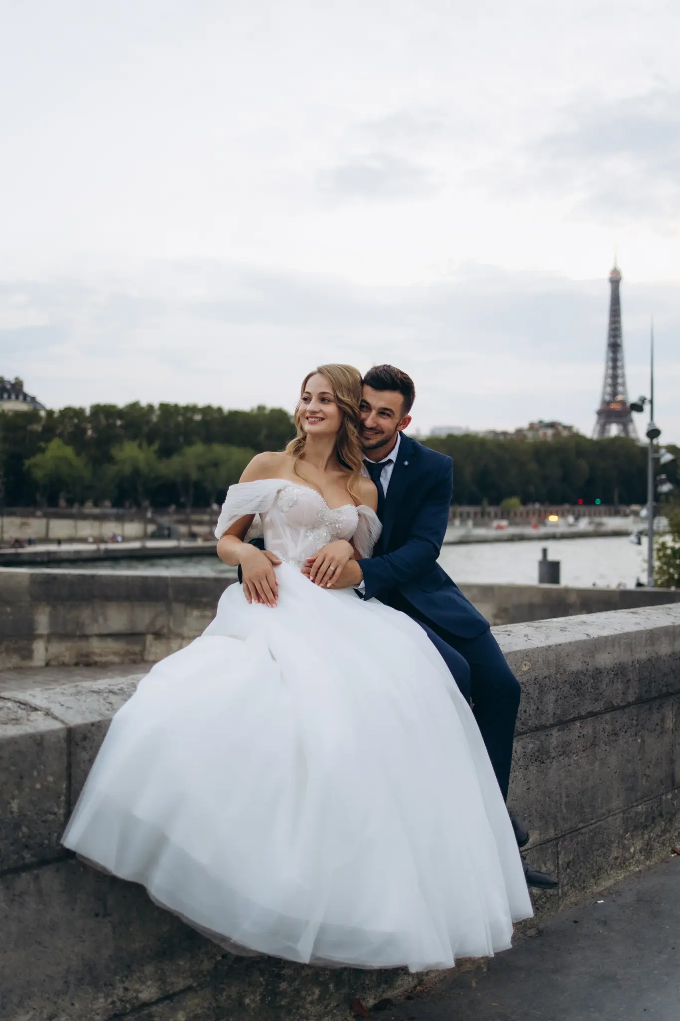 Bride sitting on the Seine wall in the groom’s arms, Eiffel Tower in the distance