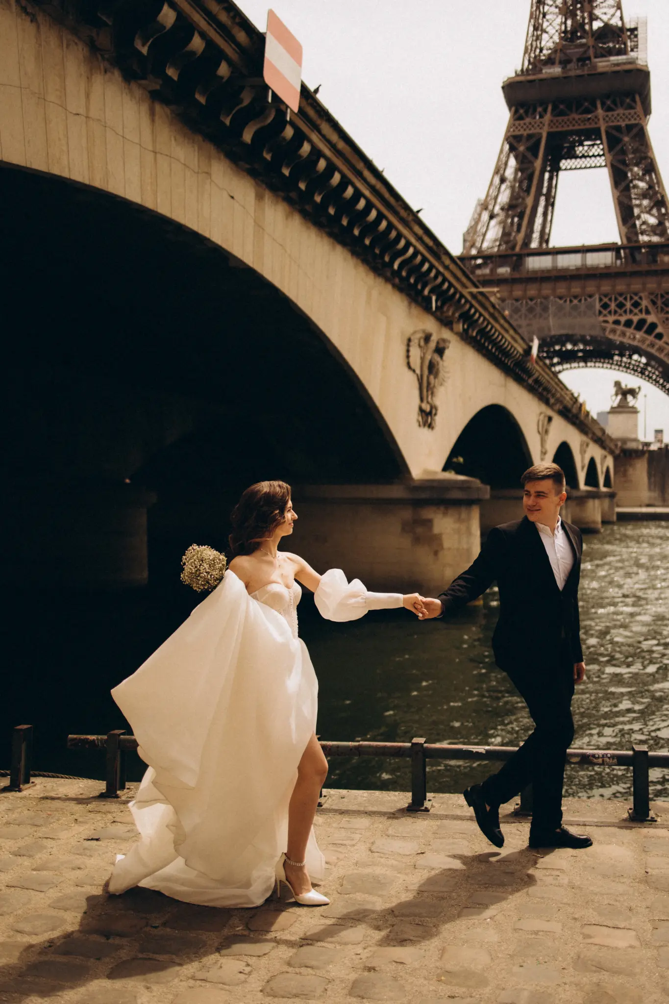 Bride with a bouquet holding the groom’s hand under the arches of Pont d’Iéna