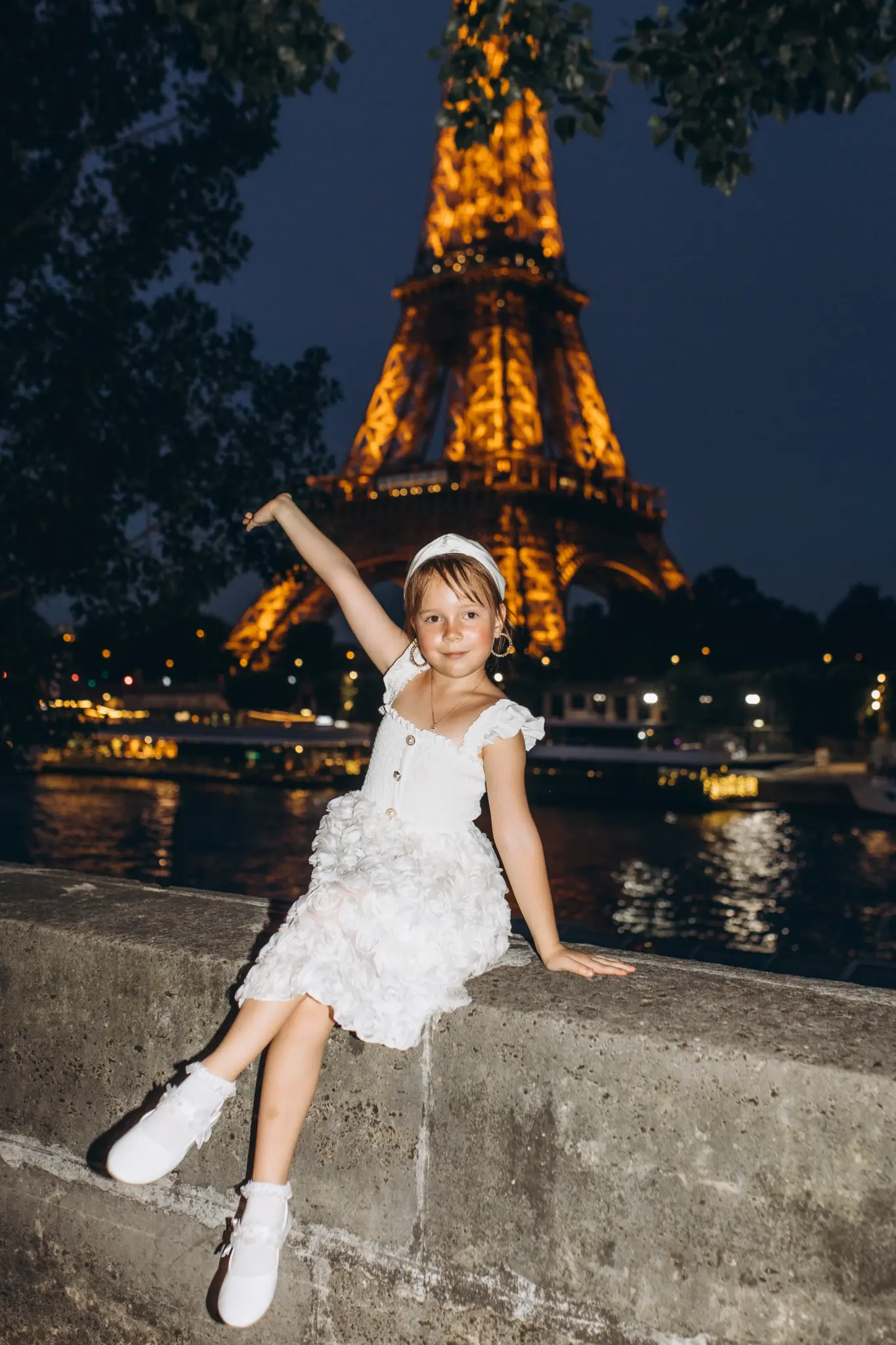 Little girl in a white dress waving on the Seine wall in front of the sparkling Eiffel Tower