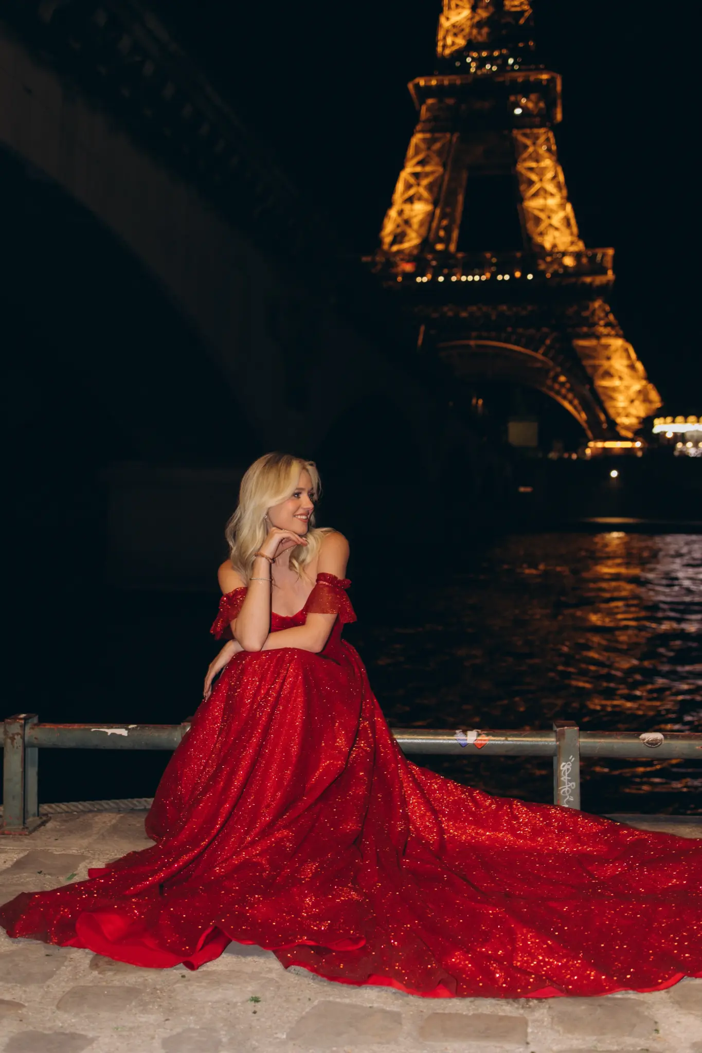Woman in a sparkling red gown with a long train by the Seine, the Eiffel Tower lit at night