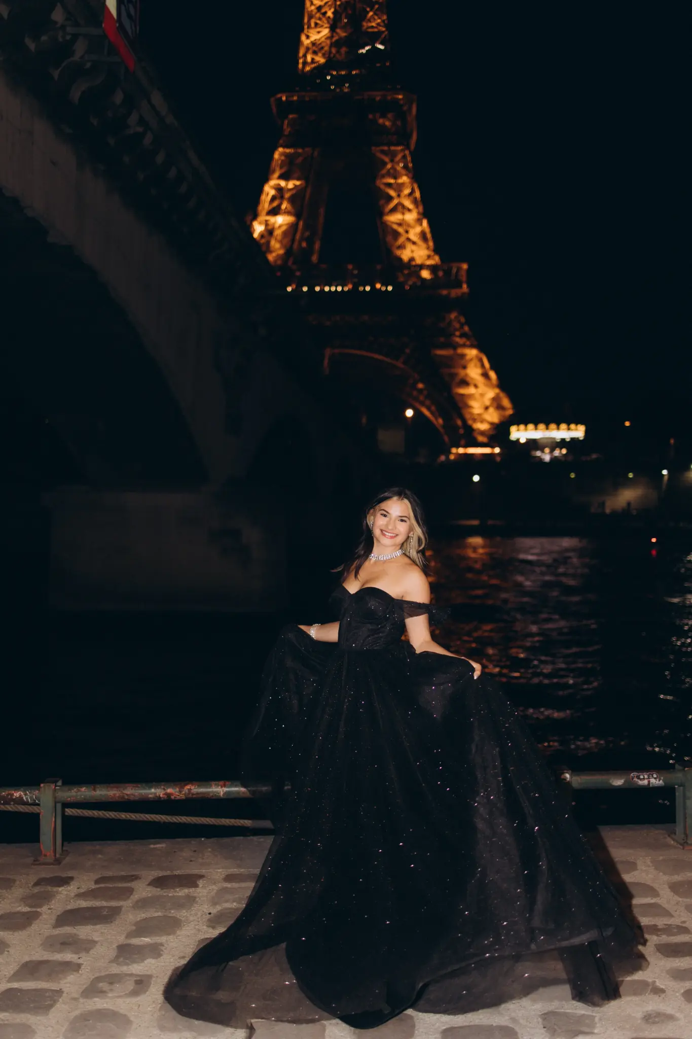 Woman in a black ball gown on the Seine bank under a bridge, the Eiffel Tower lit