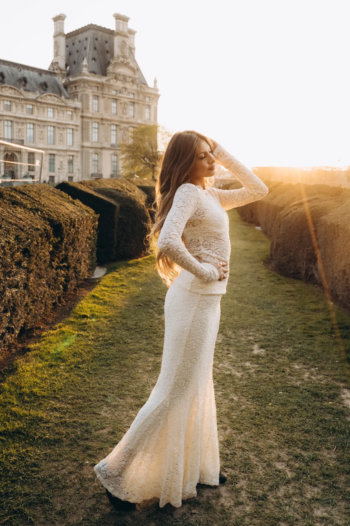 Woman in a white lace gown in the Tuileries Garden at sunset, the Louvre behind her