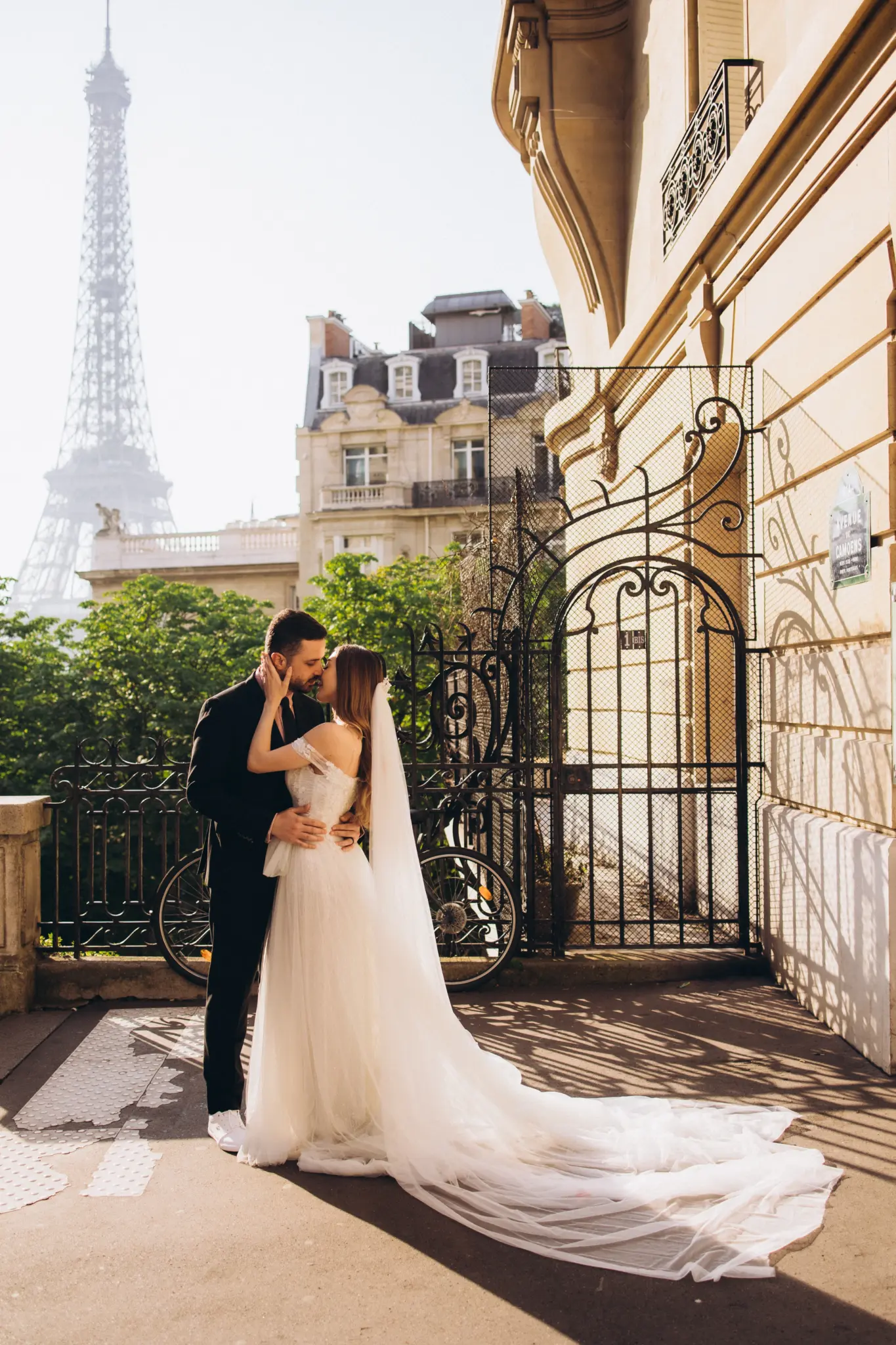 Bride with a long veil and groom by an iron gate on Avenue de Camoëns, Eiffel Tower behind them