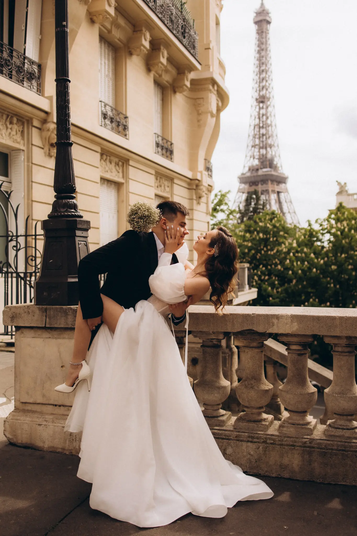 Groom lifting the bride by a stone balustrade, Haussmann building and Eiffel Tower behind