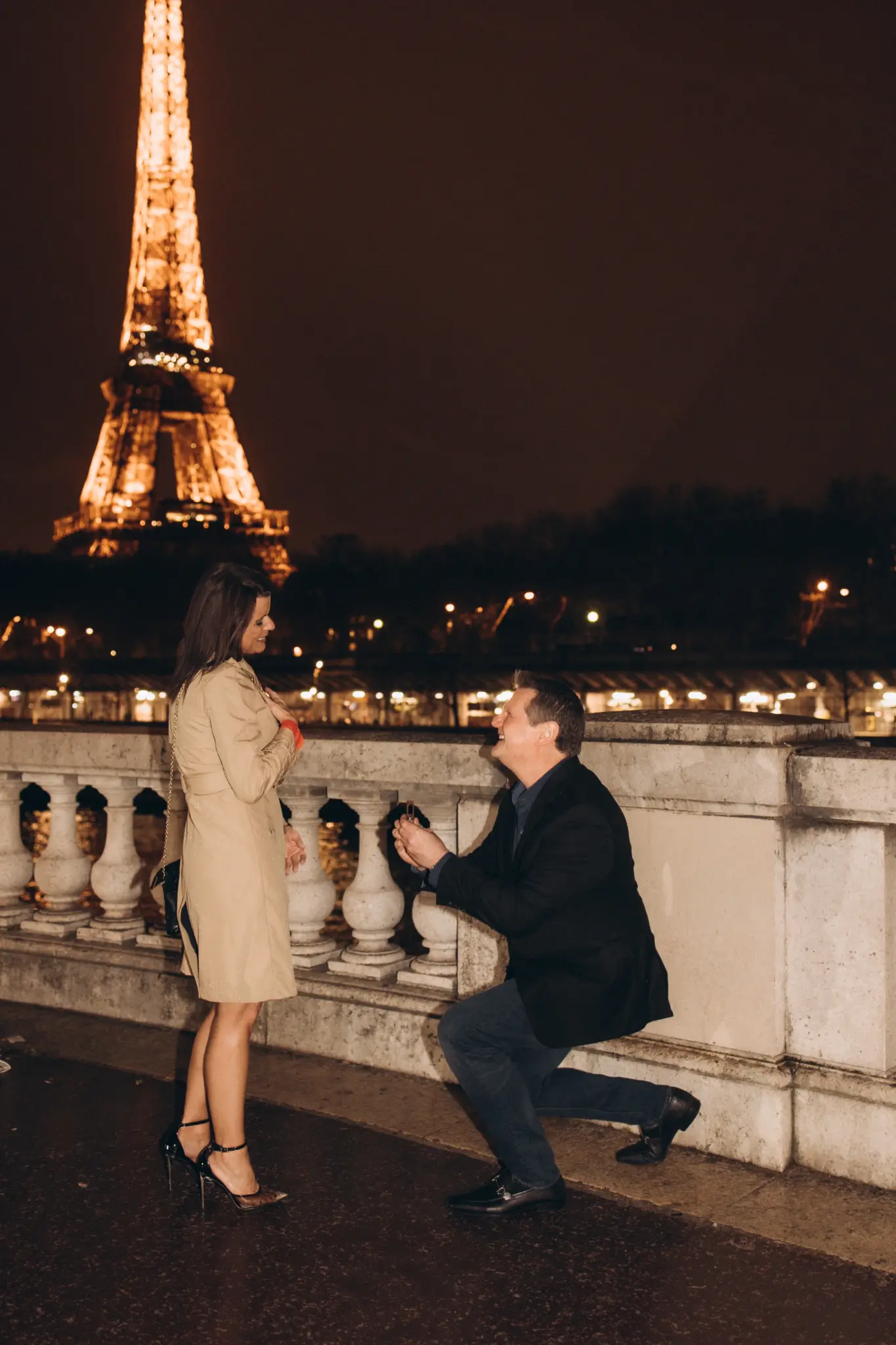 Man kneeling to propose at a stone balustrade in front of the illuminated Eiffel Tower at night