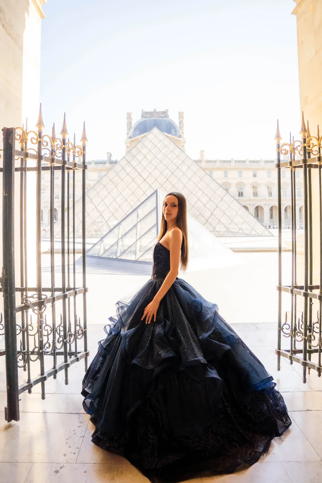 Quinceañera in a navy ball gown at an open iron gate with the Louvre pyramid behind