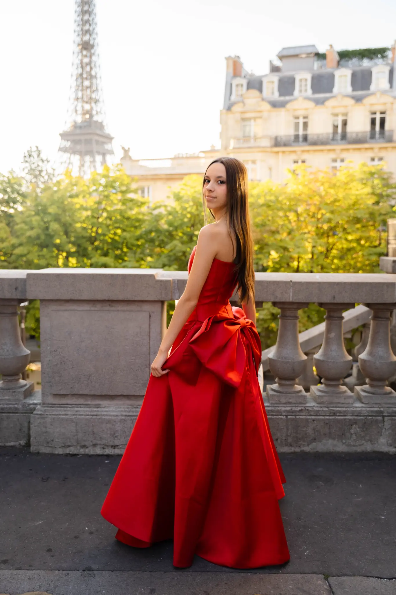 Quinceañera in a red gown with a bow at a stone balustrade near the Eiffel Tower