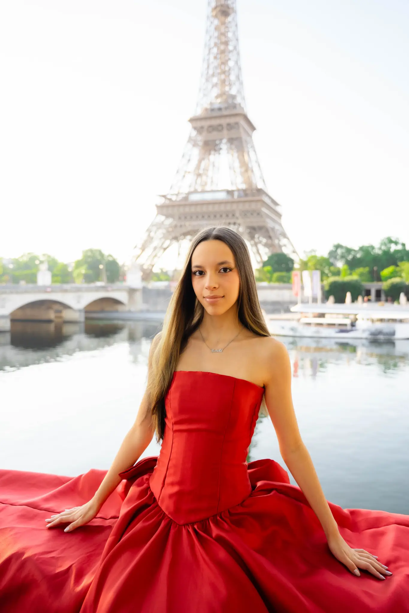 Quinceañera in a red gown sitting by the Seine with the Eiffel Tower behind