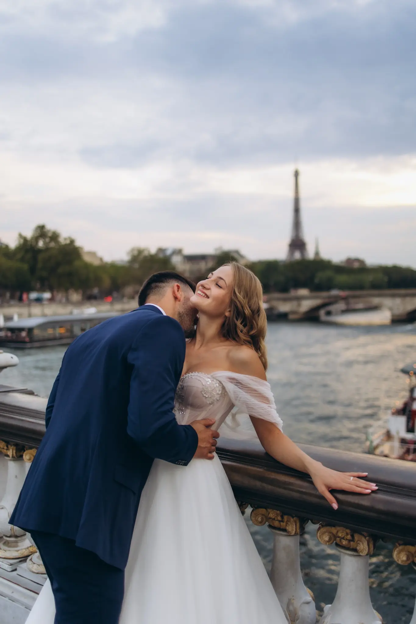 Groom kissing the bride’s cheek at the railing over the Seine, Eiffel Tower in the distance