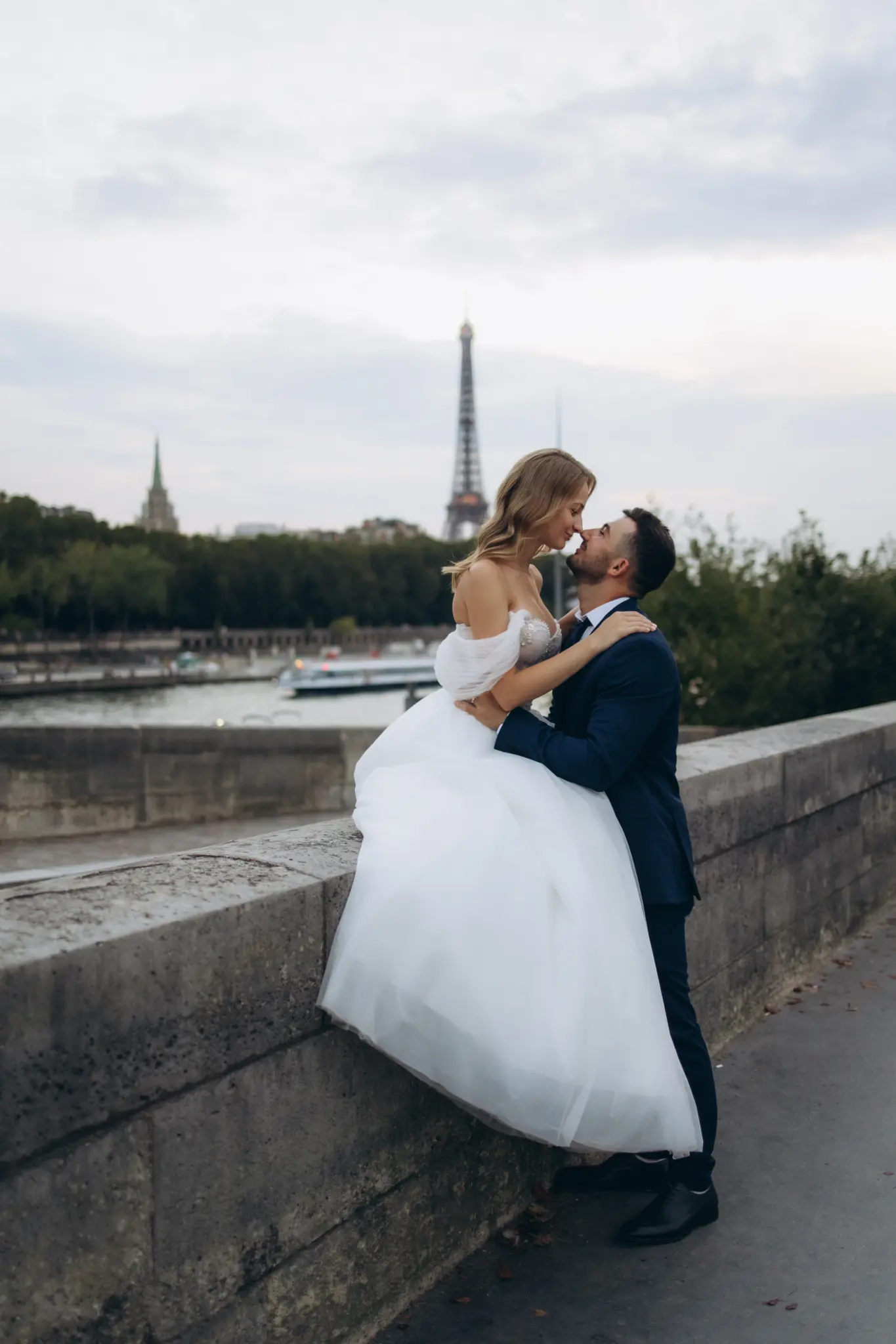 Bride sitting on the stone wall of the Seine embraced by the groom, Eiffel Tower behind