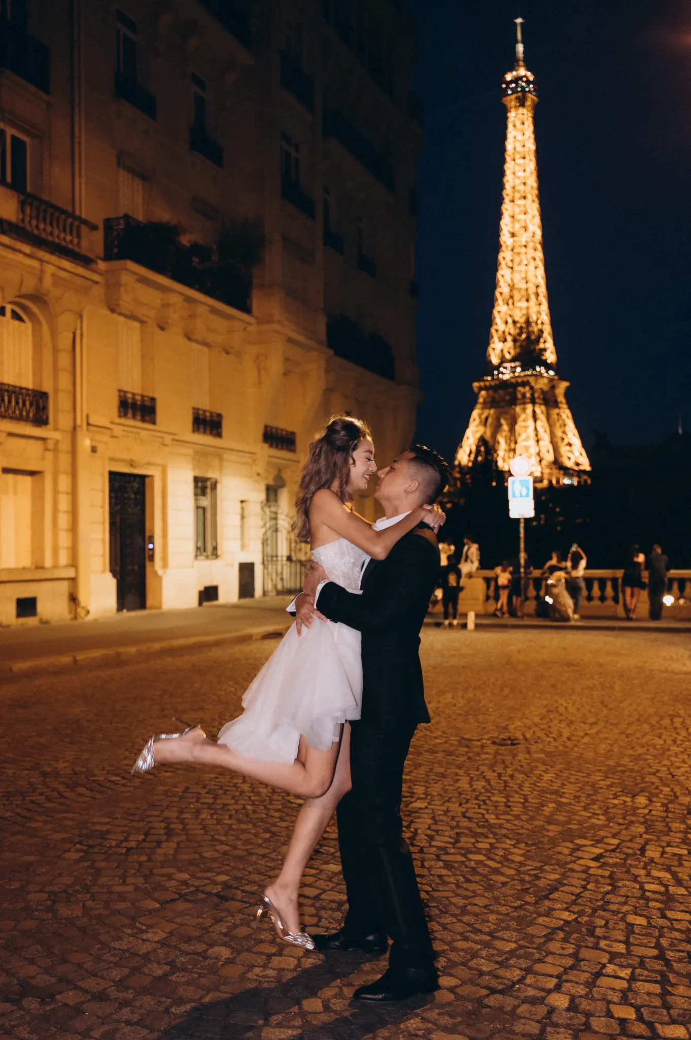 Groom lifting the bride on a cobbled street at night with the sparkling Eiffel Tower
