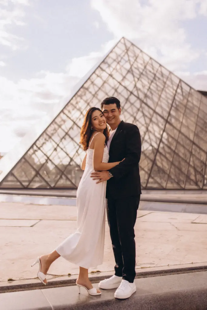Paris couple photoshoot at the Louvre, a couple in a white dress and black suit embracing in golden light before the glass pyramid