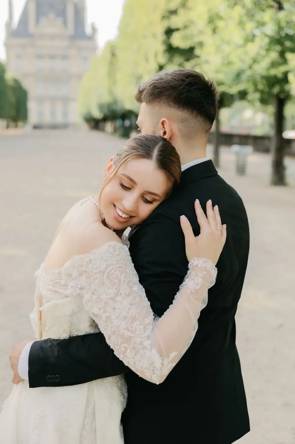 Professional couple photoshoot in the Tuileries Garden, a woman in an ivory lace gown resting her head on a man's shoulder on a tree-lined path, by Julia Litvin